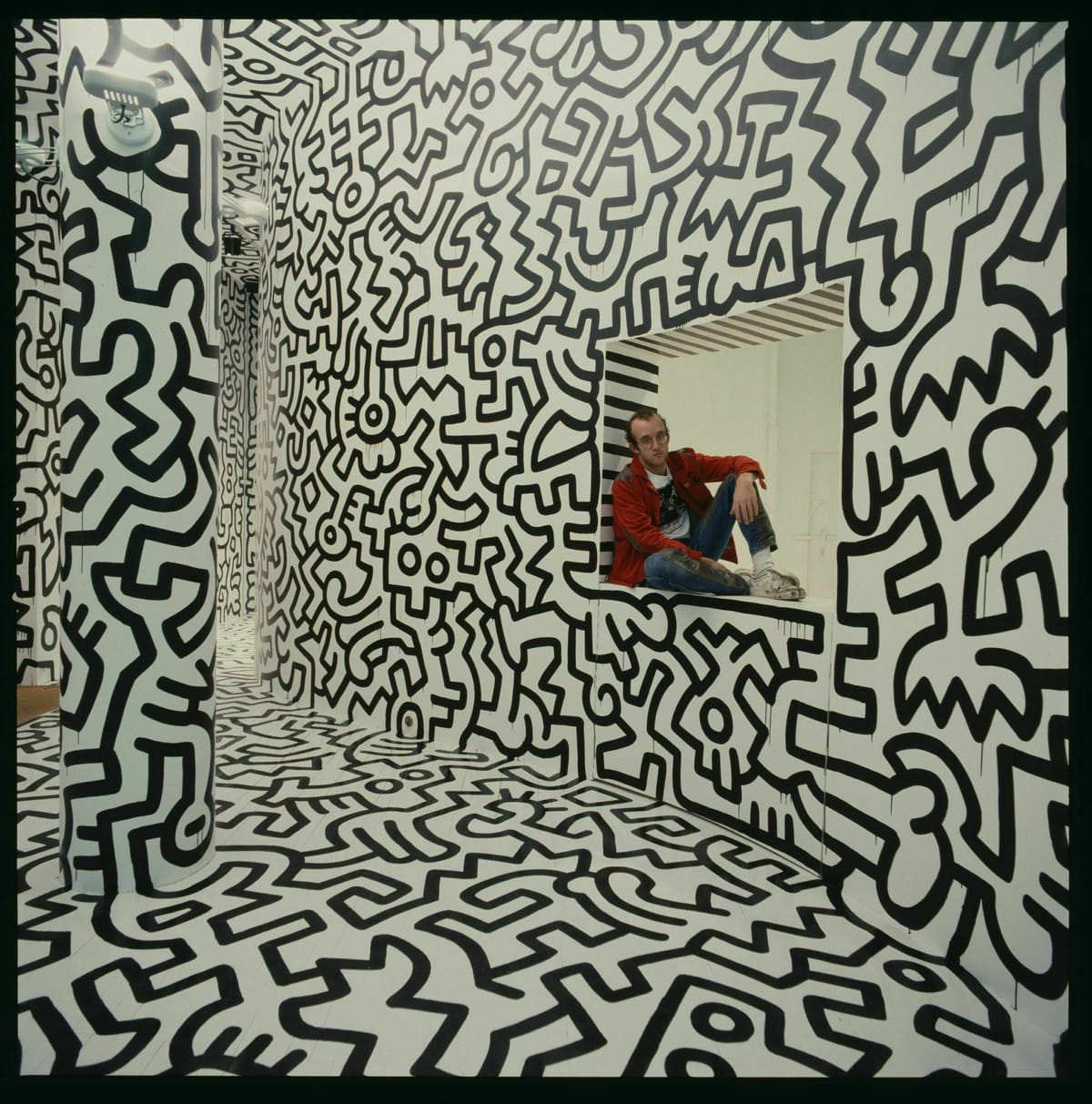 Keith Haring (May 4, 1958 – February 16, 1990) sitting amidst a giant mural he's painted.