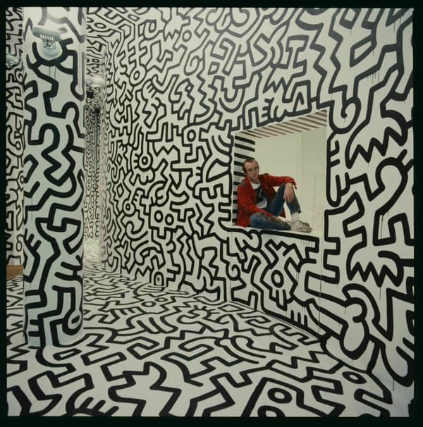 Keith Haring (May 4, 1958 – February 16, 1990) sitting amidst a giant mural he's painted. 
